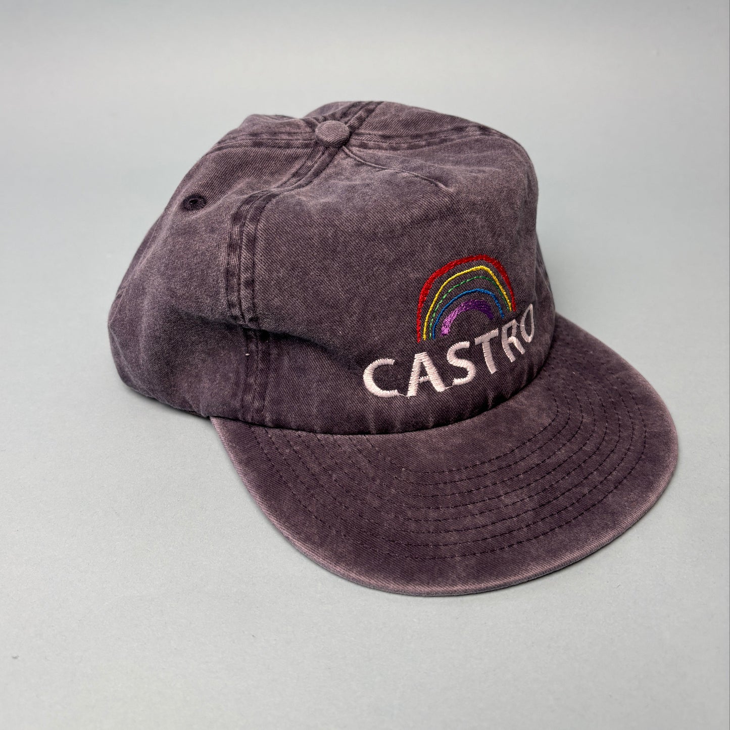 A washed dark purple baseball cap with a curved brim, featuring a colorful embroidered rainbow and white 'CASTRO' text on the side, shown against a light gray background.